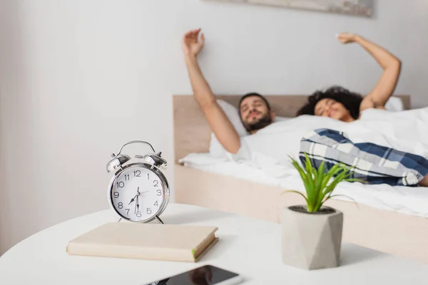 Alarm clock, book, plant and smartphone on coffee table near interracial couple on blurred background — Stock Photo