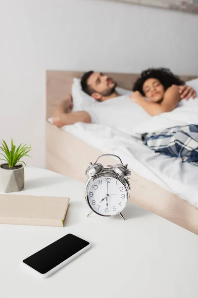 Réveil, livre, plante et smartphone avec écran blanc sur la table basse près de couple interracial sur fond flou — Photo de stock