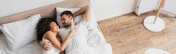 Vue du dessus de couple interracial positif étreignant et couché dans le lit, bannière — Photo de stock