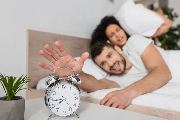 Displeased man reaching alarm clock near african american girlfriend in morning — Stock Photo