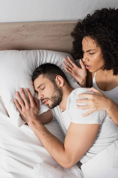 Top view of displeased african american woman looking at bearded man sleeping on bed — Stock Photo