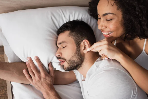 Вид зверху щаслива афроамериканська жінка торкається вуха бородатого чоловіка, який спить на ліжку — стокове фото