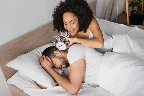 Feliz afroamericana mujer sosteniendo despertador cerca disgustado hombre durmiendo en la cama - foto de stock