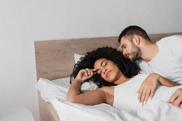 Bearded man looking at curly african american girlfriend sleeping in bed — Stock Photo