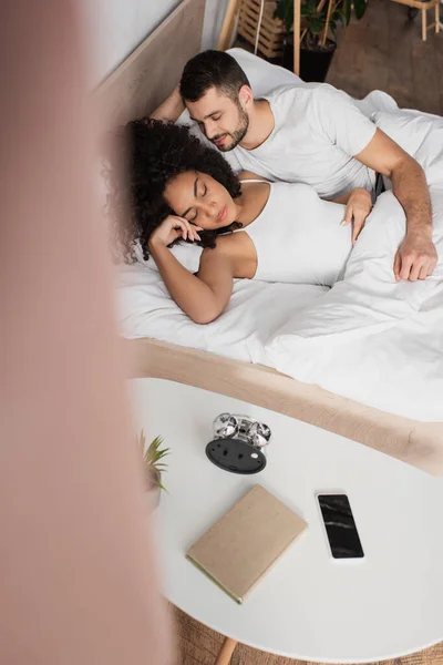 Bearded man looking at african american girlfriend sleeping in bedroom with blurred foreground — Stock Photo
