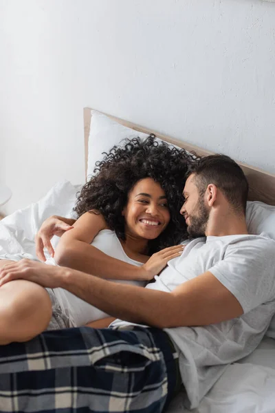 Fröhliches interrassisches Paar lächelt, während es auf dem Bett liegt — Stockfoto