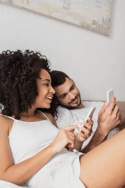 Alegre pareja interracial utilizando teléfonos celulares en el dormitorio - foto de stock