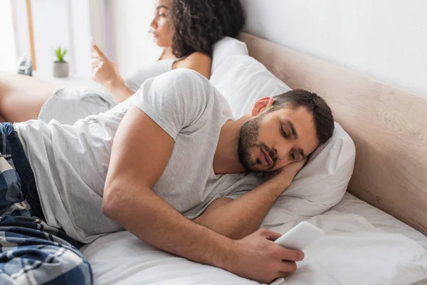 Homme barbu couché sur le lit et textos sur smartphone près de femme afro-américaine sur fond flou — Photo de stock