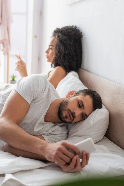 Homem barbudo deitado na cama e conversando no smartphone perto de mulher americana africana no fundo borrado — Fotografia de Stock