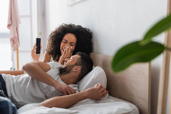 Afro-américaine femme riant et regardant petit ami tout en tenant smartphone avec écran blanc dans la chambre à coucher avec avant-plan flou — Photo de stock