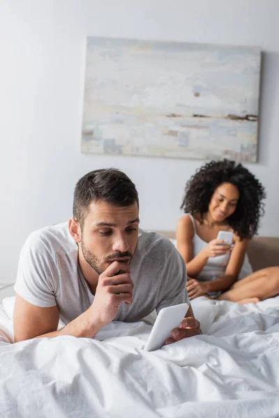 Hombre barbudo acostado en la cama y mensajes de texto en el teléfono inteligente cerca de la mujer afroamericana sobre fondo borroso - foto de stock