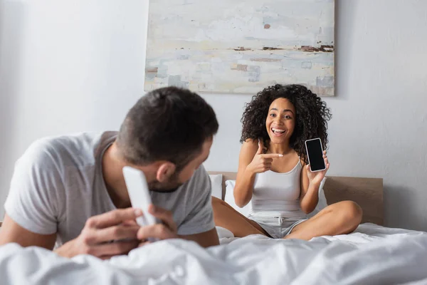 Happy african american woman pointing with finger at cellphone with blank screen while looking at boyfriend on blurred foreground — Stock Photo