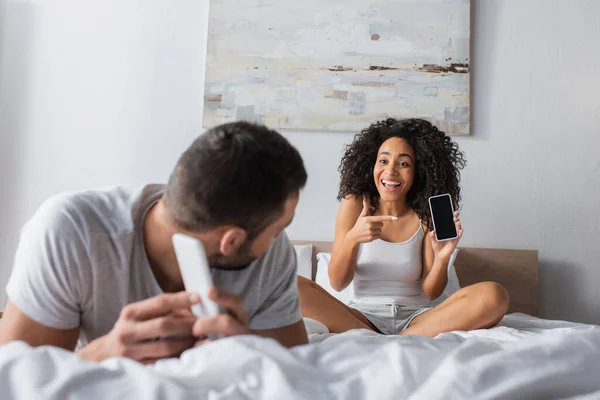 Mujer afroamericana feliz apuntando con el dedo al teléfono celular con pantalla en blanco mientras mira al novio en primer plano borroso - foto de stock