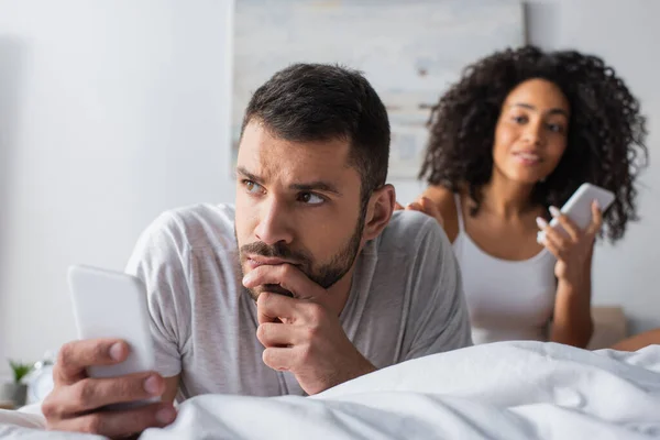 Pensive man lying on bed with smartphone near african american woman on blurred background — Stock Photo