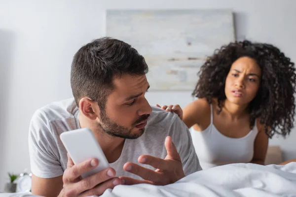 Homem descontente deitado na cama e apontando para smartphone perto de mulher americana africana em fundo borrado — Fotografia de Stock