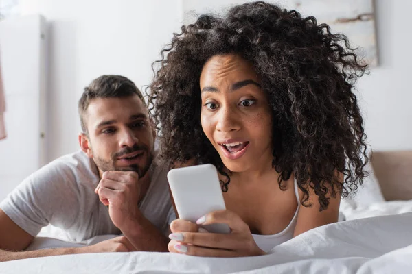 Sorprendida mujer afroamericana sosteniendo teléfono inteligente cerca novio barbudo sobre fondo borroso - foto de stock