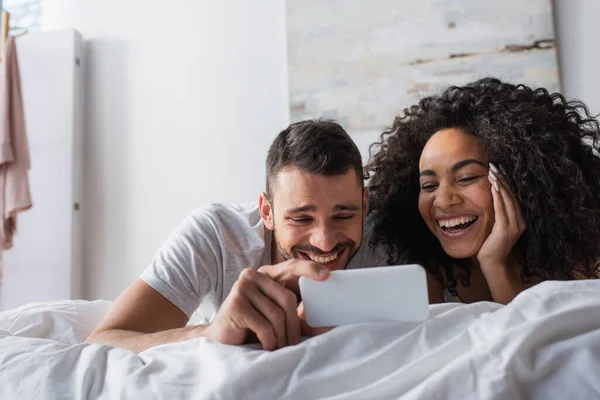 Позитивная многонациональная пара смотрит на смартфон в спальне — стоковое фото