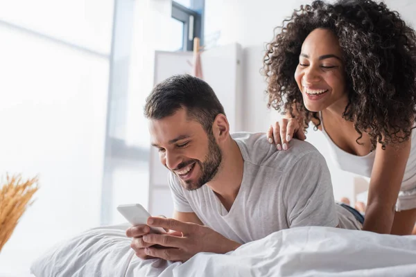 Alegre pareja multiétnica mirando teléfono inteligente en el dormitorio - foto de stock