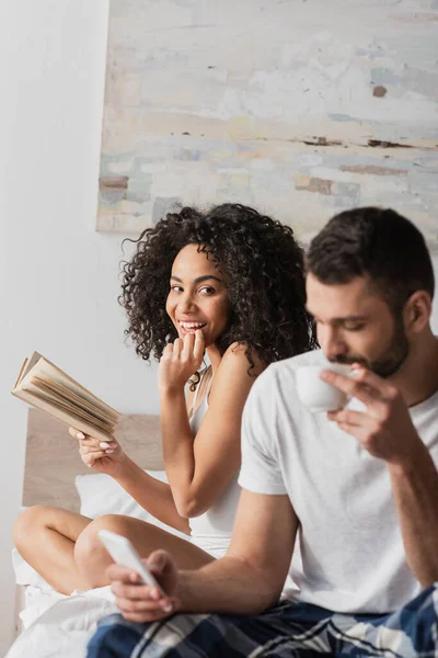 Cheerful african american woman holding book and looking at boyfriend holding smartphone while drinking coffee on blurred foreground — Stock Photo