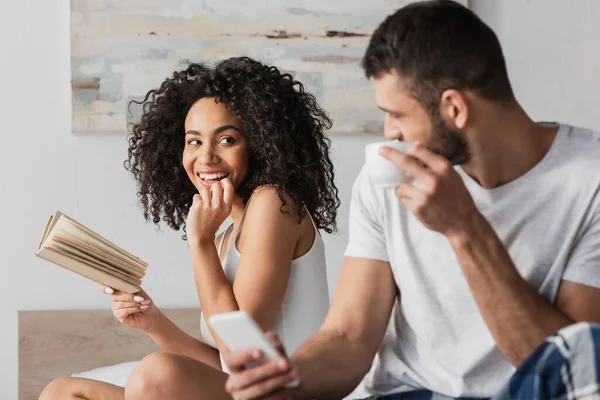 Sonriente afroamericana mujer sosteniendo libro y mirando novio sosteniendo teléfono inteligente mientras bebe café en primer plano borrosa - foto de stock