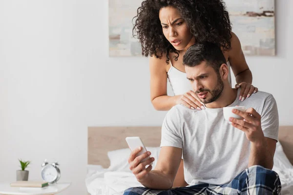 Barbudo hombre mirando el teléfono inteligente y sosteniendo la taza de café cerca sorprendido afroamericano novia - foto de stock