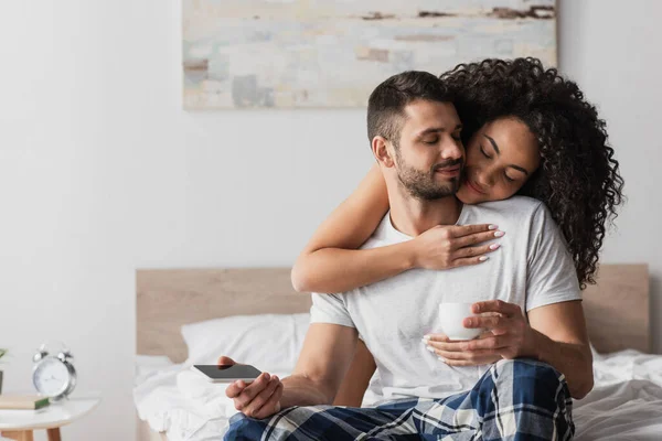 Happy african american woman hugging bearded boyfriend with smartphone and cup — Stock Photo