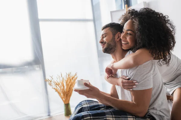 Happy african american woman hugging bearded boyfriend with cup of coffee — Stock Photo