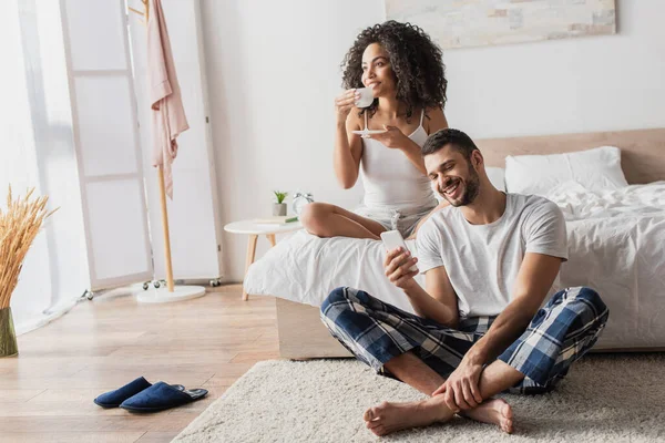 Mujer afroamericana feliz sosteniendo taza con café mientras novio barbudo usando teléfono inteligente y sentado en la alfombra - foto de stock
