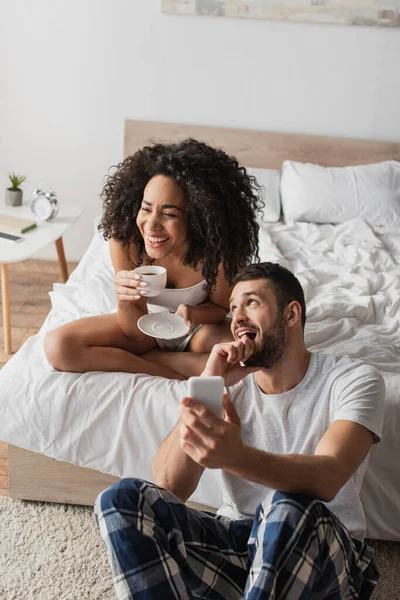 Mujer afroamericana feliz sosteniendo taza con café mientras novio barbudo usando teléfono inteligente en el dormitorio - foto de stock