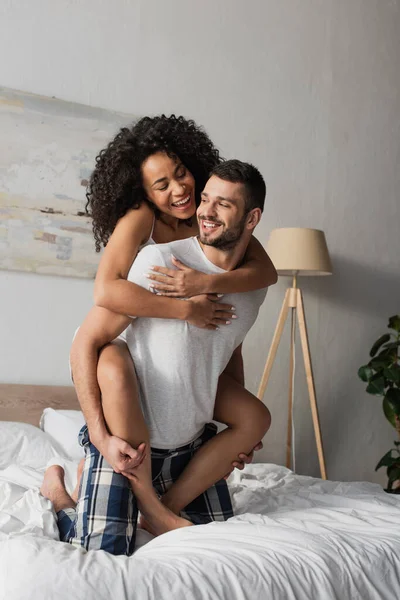 Alegre barbudo homem piggybacking afro-americano namorada no quarto — Fotografia de Stock