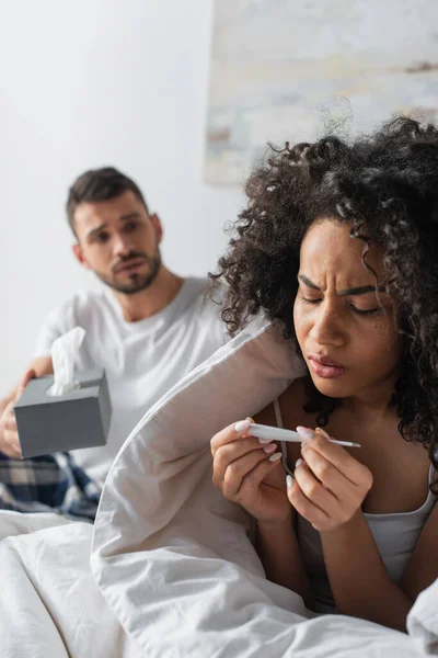 Doente Africano americano mulher olhando para o termômetro digital perto preocupado namorado no fundo borrado — Fotografia de Stock