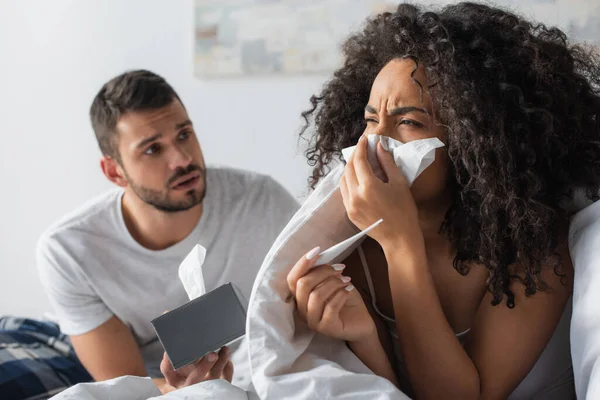 Enferma africana americana mujer estornudando en servilleta cerca preocupado novio - foto de stock