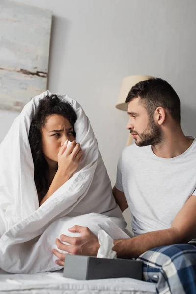 Sick african american woman under blanket sneezing in napkin near worried boyfriend — Stock Photo