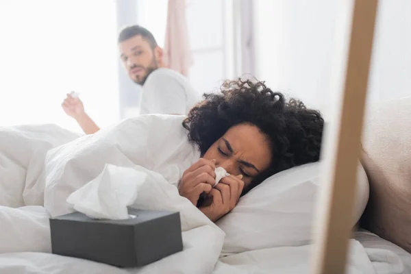 Malade afro-américaine couché sous la couverture et éternuer dans la serviette près de copain inquiet sur fond flou — Photo de stock