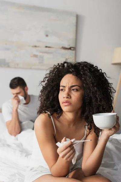 Kranke Afroamerikanerin hält digitales Thermometer und Tasse in der Nähe ihres Freundes auf verschwommenem Hintergrund — Stockfoto