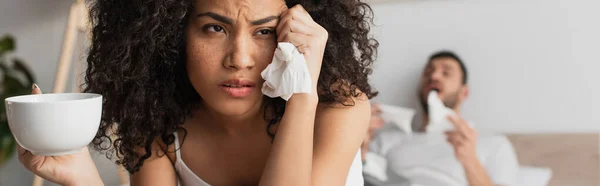 Kranke Afroamerikanerin hält Gewebe und Tasse in der Nähe ihres Freundes auf verschwommenem Hintergrund, Banner — Stockfoto