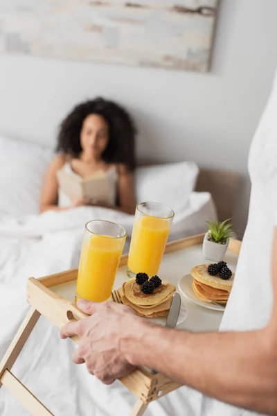 Visão parcial do homem segurando bandeja com saboroso café da manhã perto feliz afro-americana namorada no fundo borrado — Fotografia de Stock