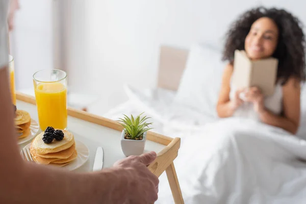 Abgeschnittene Ansicht des Mannes hält Tablett mit leckerem Frühstück in der Nähe glücklich afrikanisch-amerikanische Freundin auf verschwommenem Hintergrund — Stockfoto