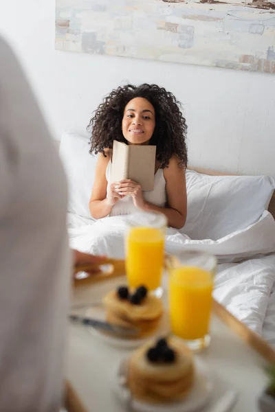 Homem desfocado segurando bandeja com café da manhã perto feliz afro-americana namorada com livro — Fotografia de Stock