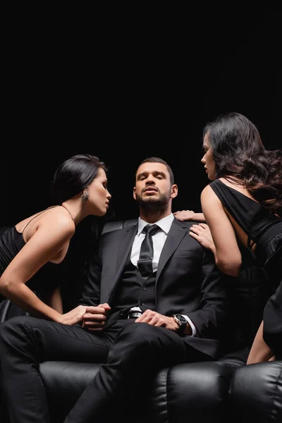 Successful businessman sitting on leather couch near women seducing him isolated on black — Stock Photo