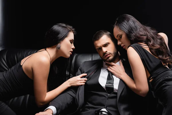 Empresário de sucesso perto de mulheres apaixonadas seduzindo-o no sofá de couro em preto — Fotografia de Stock