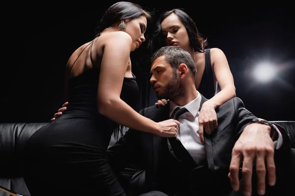 Femmes sexy déshabiller homme d'affaires assis sur le canapé en cuir sur noir — Stock Photo