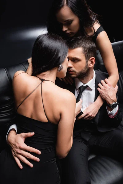 Vue grand angle de sexy, brune femmes séduire homme d'affaires sur canapé en cuir sur noir — Photo de stock