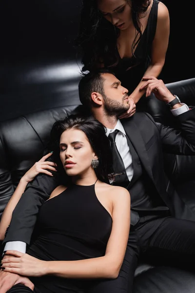 Sexy woman touching face of man sitting on leather couch on black — Stock Photo