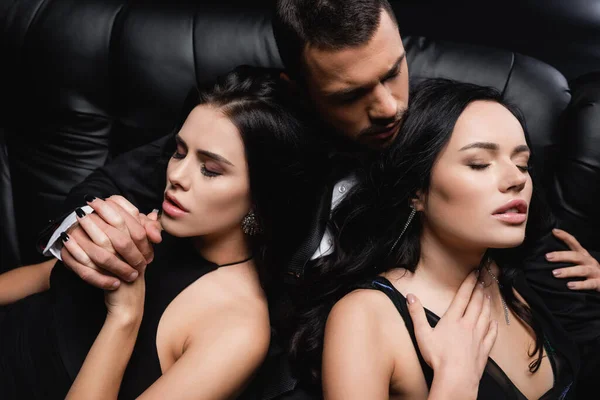 Young businessman embracing sexy women while sitting on leather couch on black — Stock Photo