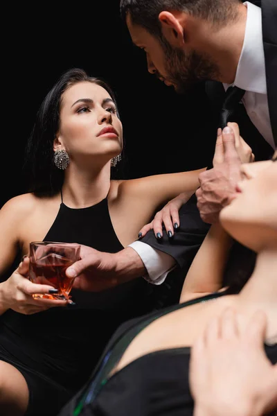 Uomo che tiene un bicchiere di whisky vicino a donne appassionate in primo piano sfocato isolato sul nero — Foto stock