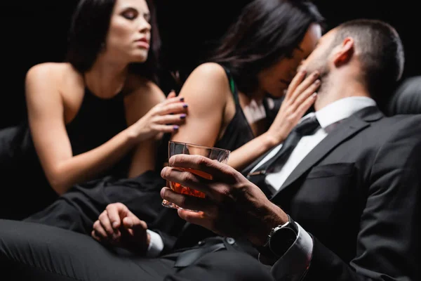 Hombre sosteniendo vaso de whisky cerca de mujeres apasionadas seduciéndolo aislado en negro, fondo borroso - foto de stock
