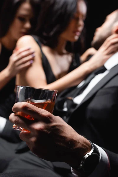 Homme avec verre de whisky près de femmes passionnées sur fond flou isolé sur noir — Photo de stock