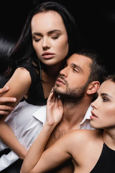 Leidenschaftliche Frauen berühren Gesicht des Mannes und umarmen ihn isoliert auf schwarz — Stockfoto