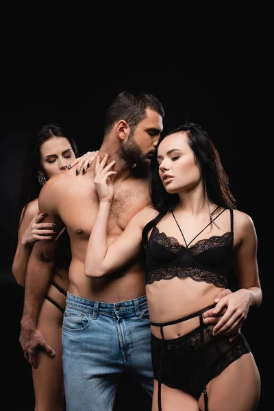 Mulheres apaixonadas em lingerie seduzindo homem sem camisa isolado em preto — Fotografia de Stock